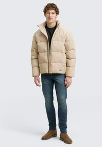 Joven con chaqueta acolchada beige, camiseta negra, jeans ajustados azules y botas de ante marrones, de pie contra un fondo gris claro.