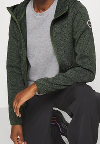 Grüne, strukturierte Reißverschlussjacke mit Kapuze, kombiniert mit einem grauen Hemd. Auffällige Details sind elastische Bündchen und ein Logo auf der Schulter.