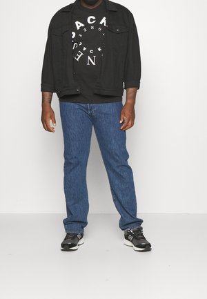 Svart denimjacka över en svart grafisk t-shirt med vit cirkulär text, blå denimjeans och svarta löparskor med vita accenter.