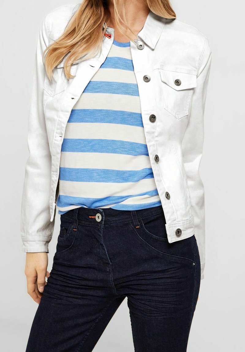 Chaqueta de mezclilla blanca con botones de metal, un bolsillo en el pecho, superpuesta sobre una camisa de rayas azules y blancas, combinada con jeans de mezclilla oscura.