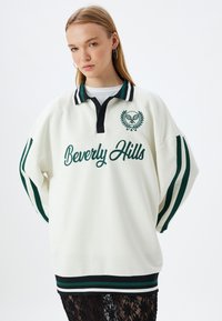 Felpa polo bianca oversize con colletto nero, strisce verdi e nere sulle maniche, con la scritta "Beverly Hills" in lettere verdi e logo.