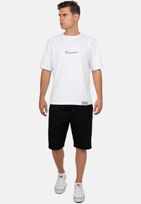 T-shirt en coton blanc à manches courtes avec un logo noir sur la poitrine, associé à un short noir et des baskets en toile blanches.