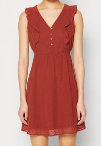 Sukienka bez rękawów w rdzawoczerwonym kolorze z fakturowanymi kropkami, falbaniastymi detalami na ramionach, dekoltem w serek oraz pięcioma guzikami z przodu, noszona przez modelkę.
