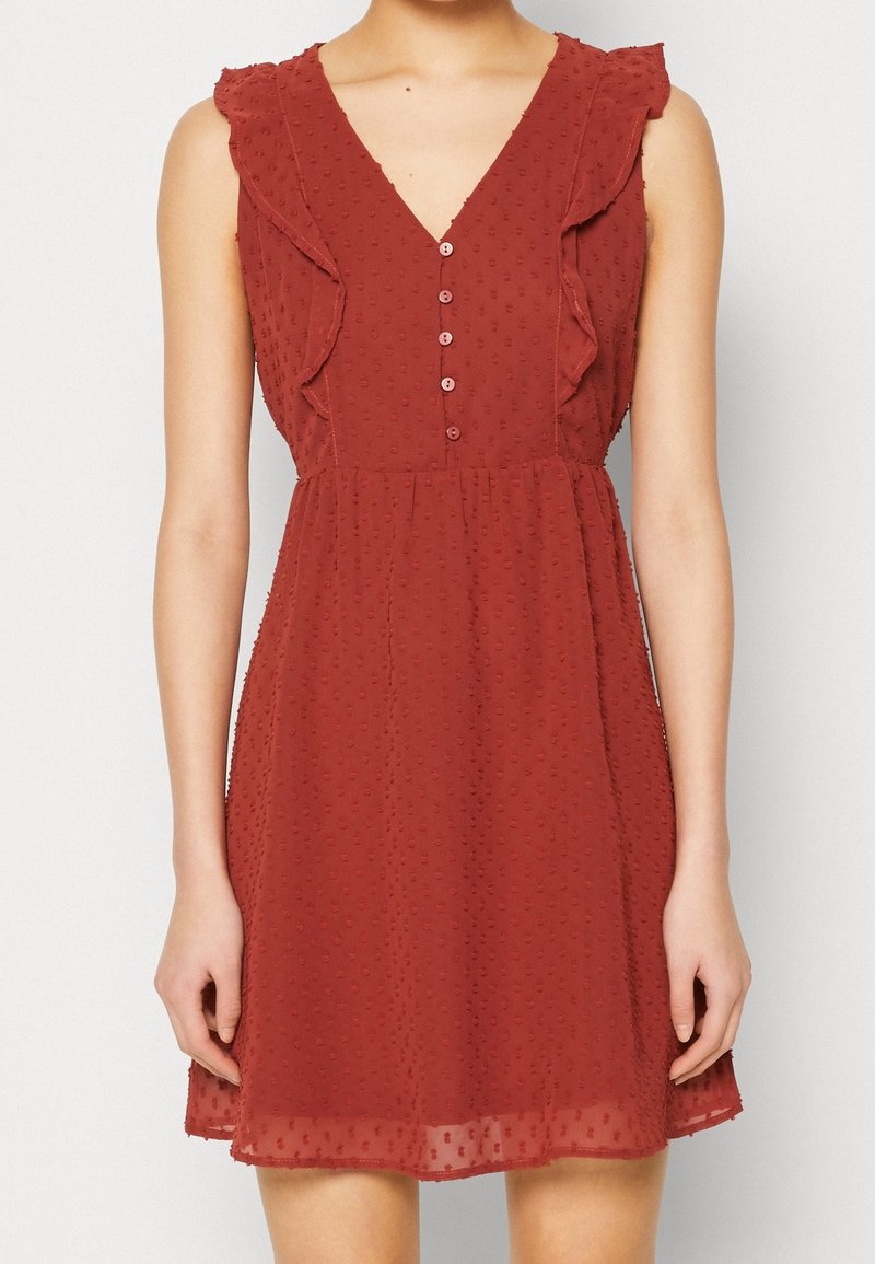 Sukienka bez rękawów w rdzawoczerwonym kolorze z fakturowanymi kropkami, falbaniastymi detalami na ramionach, dekoltem w serek oraz pięcioma guzikami z przodu, noszona przez modelkę.