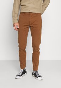Chinos en coton marron à coupe slim, avec une fermeture standard par bouton et des passants pour ceinture, assortis à des baskets en toile noire.