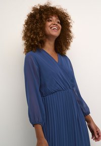 Long, deep blue dress with pleated skirt, V-neckline, and sheer long sleeves. Soft, flowing fabric adds texture. No visible patterns or accents.