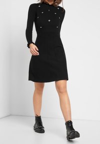 Femme portant une robe noire à manches longues ornée de petites abeilles argentées sur le haut, et des bottines noires à lacets avec des chaînes décoratives.