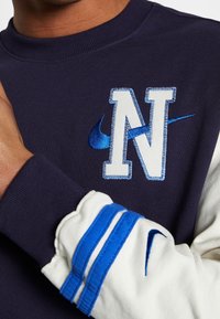 Navy sweatshirt with beige sleeves, featuring an embroidered blue "N" and Nike swoosh. Ribbed cuffs with blue stripes.