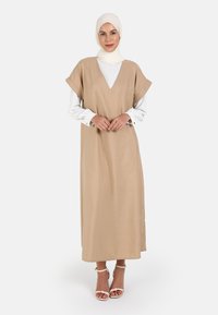 Beige maxi šaty s véčkovým výstřihem, krátkými rukávy a hladkou texturou, doplněné bílými dlouhými rukávy a sandály na pásku.