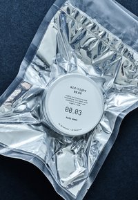 Hårmaske i en slank sølvpose med en rund hvit beholder inni, merket "mid/night 00.00" og "00.03", med minimal tekst.