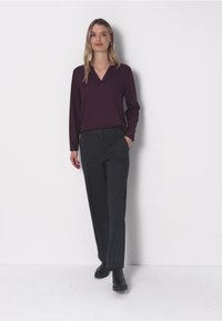 Frau steht vor weißem Hintergrund und trägt eine dunkelviolette langärmelige Bluse, schwarze Hose und schwarze Stiefeletten, mit einer Hand in der Tasche.