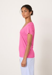 Roza majica z V-izrezom in kratkimi rokavi, izdelana iz mehke tkanine, z rahlo prilego in subtilno teksturo, nošena z svetlo modrimi hlačami z navpičnimi črtami.