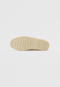 Espadrillsoffa i beige med en texturerad yta, med horisontella ränder och diskret logga på sidan. Ren design utan mönster.
