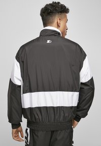 Jachetă în stil windbreaker alb-negru, cu guler înalt, manșete elastice și talie bandată. Detalii ale logo-ului vizibile pe spate.