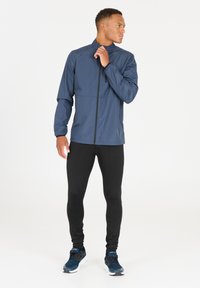Marineblauwe lichte jas met rits aan de voorkant, staatkraag en elastische manchetten, gecombineerd met strakke zwarte leggings en blauwe sportschoenen.