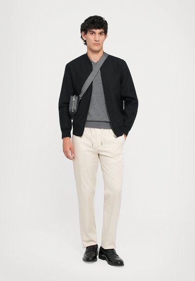 Jachetă bomber neagră, tricou gri, pantaloni crem; modelul poartă pantofi negri și ține o geantă mică gri de umăr, fundal simplu.