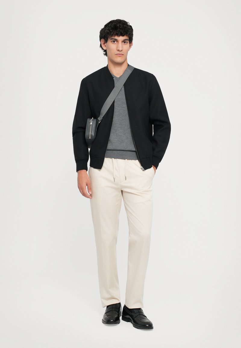 Zwarte bomberjack, grijs t-shirt, crèmekleurige broeken; model draagt zwarte schoenen en heeft een kleine grijze crossbody tas, effen achtergrond.