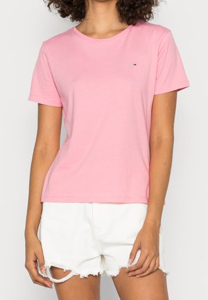 Vrouw draagt een effen roze T-shirt met korte mouwen en witte gescheurde denim shorts, staand tegen een lichte achtergrond.