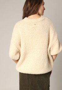 Cr ème kleurige gebreide trui met een losse pasvorm, verlaagde schouders en korte mouwen. Zachte textuur en geribde details bij de hals.