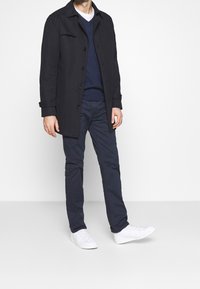 Tmavě navy kabát s knoflíky, vrstvený přes navy svetr, spárovaný s tmavými kalhotami a bílými teniskami. Oblečení má čistý, minimalistický design.