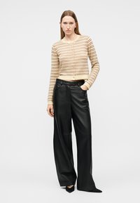 Donna che indossa un cardigan a righe beige e marrone, pantaloni in pelle neri a gamba larga e scarpe nere con punta, in piedi con una mano in tasca.