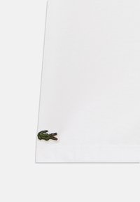 Λευκό βαμβακερό μπλουζάκι με πράσινο κεντητό λογότυπο κροκοδείλου στο κάτω αριστερό μέρος, που αναδεικνύει την λεπτή ραφή και την ομαλή υφή.