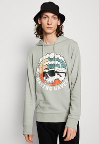 Felpa verde chiaro con un design grafico che presenta un teschio e onde. Dispone di una tasca a canguro frontale e cappuccio con cordino. Cappello a secchiello nero.