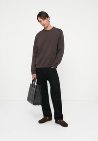 Sudadera marrón oscuro de mangas largas, pantalones negros y bolsa de cuero negra. El modelo posa sobre un fondo blanco.
