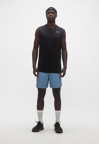 Άνδρας στέκεται φορώντας μαύρο αμάνικο μπλουζάκι Nike, μπλε σορτς, λευκές κάλτσες μέσης, μαύρα αθλητικά παπούτσια και μαύρη κορδέλα Nike στο κεφάλι, μπροστά σε λευκό φόντο.