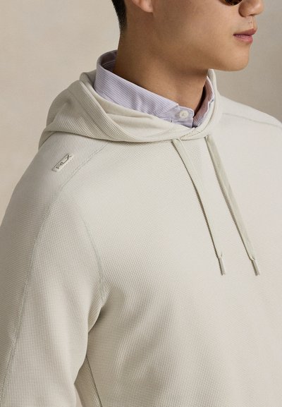 Ανοιχτό μπεζ φούτερ με υφή υφάσματος, κορδόνια και τσέπη καγκουρό. Διακοσμημένο με το λογότυπο RLX στον ώμο. Φορεμένο πάνω από πουκάμισο με γιακά.