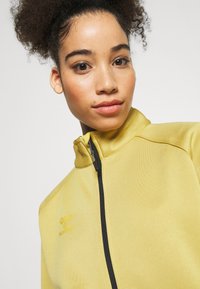 Żółta kurtka zapinana na zamek z wysokim kołnierzem, teksturowana tkanina i czarny zamek. Posiada logo na lewej stronie klatki piersiowej.