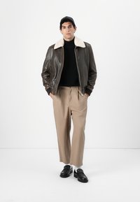 Hnedá kožená bomber bunda s bielym ovčím golierom, čierny rolák, béžové nohavice a čierne poltopánky na modeli na bielom pozadí.