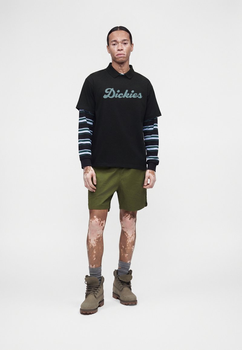 Junger Mann mit Vitiligo, der ein schwarzes Dickies Poloshirt über einem gestreiften Langarmshirt, grüne Shorts, graue Socken und braune Stiefel trägt.