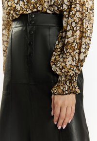Zwarte leren rok met een hoge taille en knoopdetails. Vergezeld van een bloemige, transparante blouse met lange mouwen en ruches bij de manchetten.