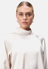 Svetlo béžový top s krátkym golierom vyrobený z hladkej tkaniny, s raglánovými rukávmi a minimalistickým dizajnom. Jemný lesk pridáva elegantný vzhľad.
