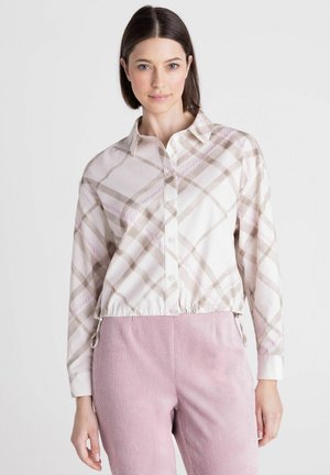 Jonge vrouw draagt een beige en roze geruit overhemd met knopen en hooggetailleerde roze corduroy broek tegen een effen witte achtergrond.