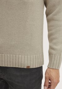 Pull en tricot beige avec un ourlet et des poignets côtelés ; présente un tissage texturé et une petite étiquette en cuir en bas à gauche.