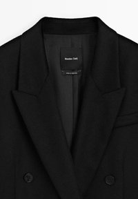 Zwarte wollen jas met een dubbelgebreid ontwerp, gekraagde kleppen, subtiele textuur en twee knopen; label leest "Massimo Dutti - Made in Portugal."