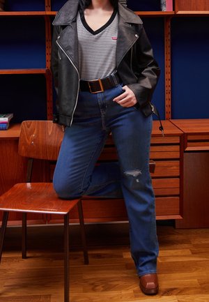 Person iført en sort læderjakke, striped skjorte, blå jeans med en revne og brune sko, der knæler med det ene ben på en træstol.