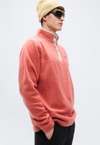 Πουλόβερ από κοραλλένιο φλις με ψηλό γιακά, κουμπιά σε αντίθετο μπεζ χρώμα και κεντημένο λογότυπο. Το μοντέλο φορά επίσης μπεζ σκούφο και γυαλιά ηλίου.