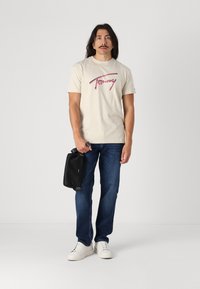 T-shirt en coton beige avec un design graphique en texte rose, associé à un jean en denim bleu et des baskets blanches. Tenant un sac à main noir.