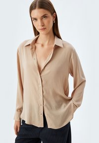Blusa beige a maniche lunghe con vestibilità comoda, chiusura con bottoni sul davanti e colletto classico. Tessuto liscio con un leggero lucido.