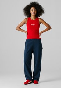 Kobieta o kręconych włosach stoi z rękami na biodrach, ubrana w czerwony tank top "Red Bull Racing", granatowe spodnie i czerwone tenisówki.