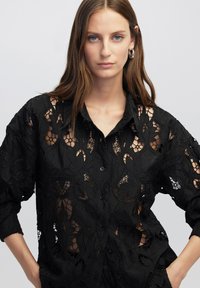 Maglietta nera con motivo in pizzo floreale e sezioni trasparenti, chiusura frontale con bottoni e maniche lunghe. Tessuto testurizzato con dettagli intricati.