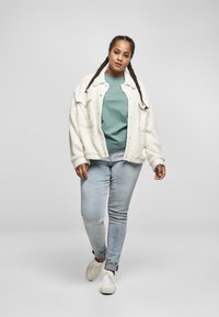 Hvit, spendt jakke over en teal skjorte, sammen med lyseblå jeans og hvite joggesko. Jakken har en løs passform og knapp detaljer.