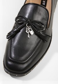 Mocassins en cuir noir avec un détail en nœud orné de pompons et d'un petit charme en forme de clé. La semelle est décorée de clous en argent. Texture lisse, design classique.