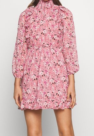 Kobieta ubrana w różową mini sukienkę w kwiaty, z długimi bufkami, falbanianym wysokim kołnierzem, talią podkreśloną paskiem i falbanianym dołem.