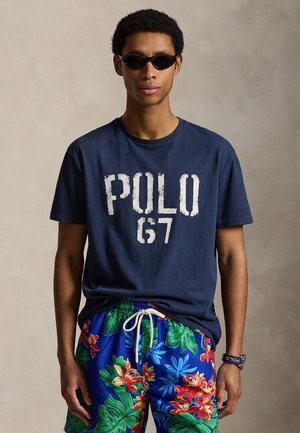 Man wearing black sunglasses, navy blue "POLO 67" t-shirt, floral blue shorts, and a wristwatch against a neutral background.