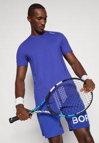 Niebieska koszulka sportowa z krótkim rękawem, teksturowana tkanina. Pasujące niebieskie szorty z białym napisem "BORG". Trzymając rakietę tenisową o czarno-niebieskim designie.