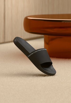 Czarny klapek Calvin Klein ustawiony pod kątem na beżowym dywanie, w tle błyszczący, zakrzywiony stół w kolorze pomarańczowo-brązowym.